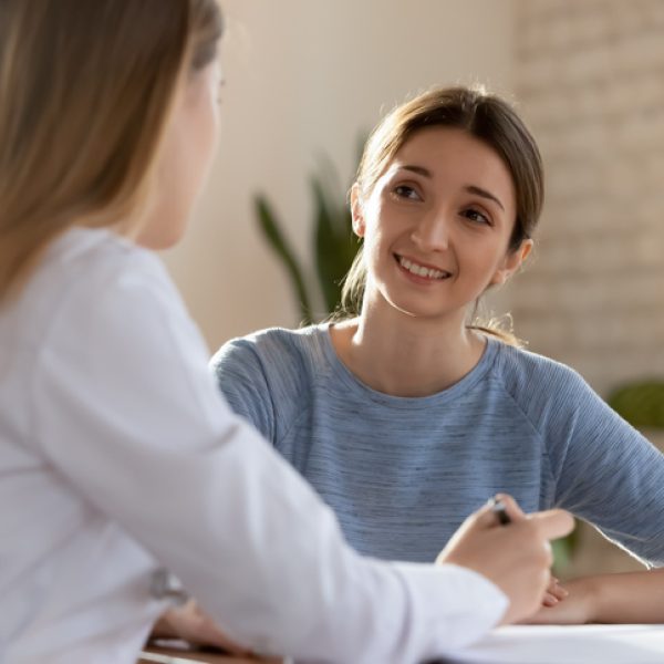Smiling millennial woman visit female doctor consulting about health complains, happy satisfied young client talk discuss treatment prescription with GP or physician at appointment in hospital